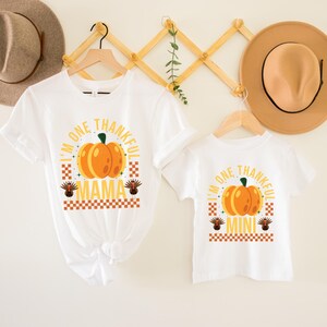 May include: Two white t-shirts with a Thanksgiving theme. The larger shirt says "I'm One Thankful Mama" with a pumpkin and a turkey. The smaller shirt says "I'm One Thankful Mini" with a pumpkin and a turkey.