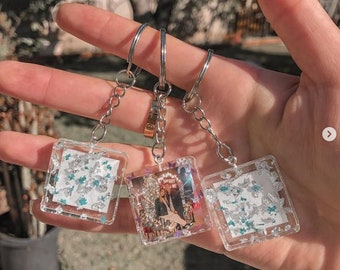 May include: Three square resin keychains with silver glitter and a clear background. The middle keychain has a photo of a couple in front of a Christmas tree. The keychains are attached to silver chains and key rings.