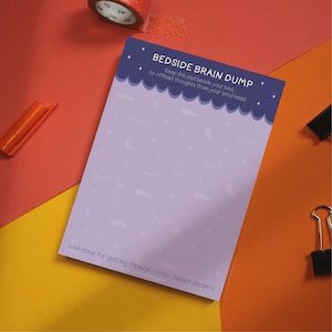 May include: A purple notepad with a blue border and white stars, clouds, and eyes. The notepad says "Bedside Brain Dump" and "Keep this pad beside your bed to offload thoughts from your tired head." The bottom of the notepad says "Well done for getting through today. Sweet dreams!"