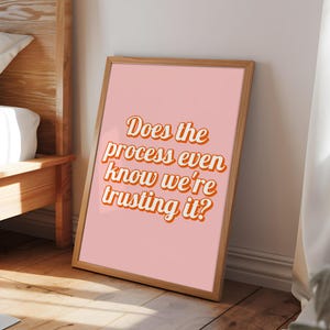 May include: A framed print with the text "Does the process even know we're trusting it?" in orange and white lettering. The print has a pink background and a wooden frame. The artwork is leaning against a wall.