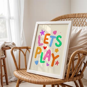 May include: A framed print with the words "LET'S PLAY" in colourful, playful lettering. The artwork features a light beige background with various shapes and colours, including stars, hearts, and flowers. The print is displayed on a wicker chair.