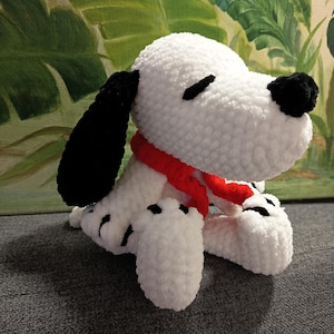 Puede incluir: Un perro de peluche blanco y negro hecho a mano con una bufanda roja. El perro tiene orejas, ojos y nariz negros. El peluche está sentado sobre una superficie gris, con un fondo pintado de verde y amarillo.