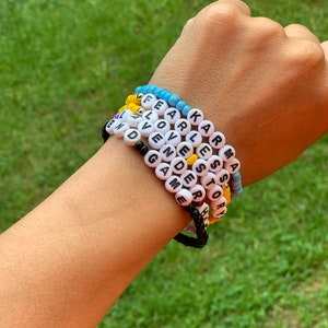 May include: A hand wearing six beaded bracelets with the words "KARMA", "STORY", "FEARLESS", "LOVE", "AVEN", and "GAME" spelled out in black letters on white beads.