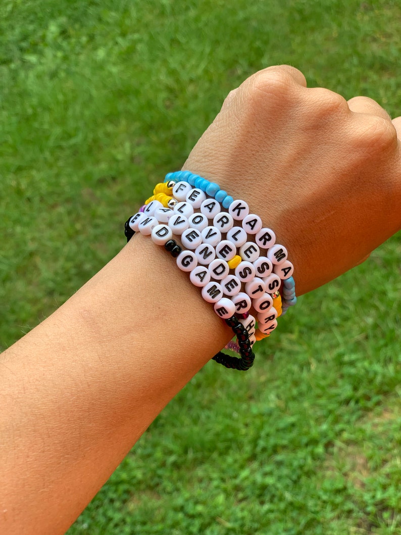 May include: A hand wearing six beaded bracelets with the words "KARMA", "STORY", "FEARLESS", "LOVE", "AVEN", and "GAME" spelled out in black letters on white beads.