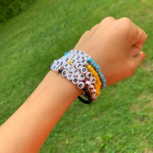 May include: A close-up of a person's wrist wearing several beaded bracelets. The bracelets spell out the words "Karma", "Yes", "Story", "End Game", "Sorry", "Amazing", and "Y'all".