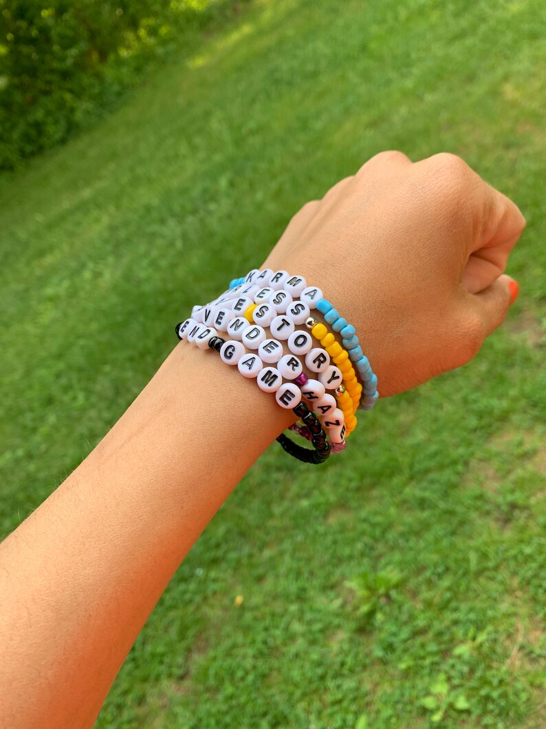 May include: A close-up of a person's wrist wearing several beaded bracelets. The bracelets spell out the words "Karma", "Yes", "Story", "End Game", "Sorry", "Amazing", and "Y'all".