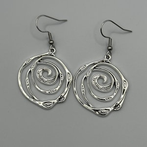 May include: A pair of silver-toned dangle earrings featuring a circular, open-work spiral design. Each earring is suspended from a standard hook wire and displays a textured, organic pattern along the swirling metal lines, creating a layered effect.