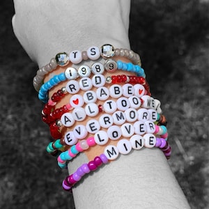 May include: A stack of colourful beaded bracelets with white letter beads spelling out the words "TS 1989 RED BABE TOO WE ALL ERMORE LOVER MINE".
