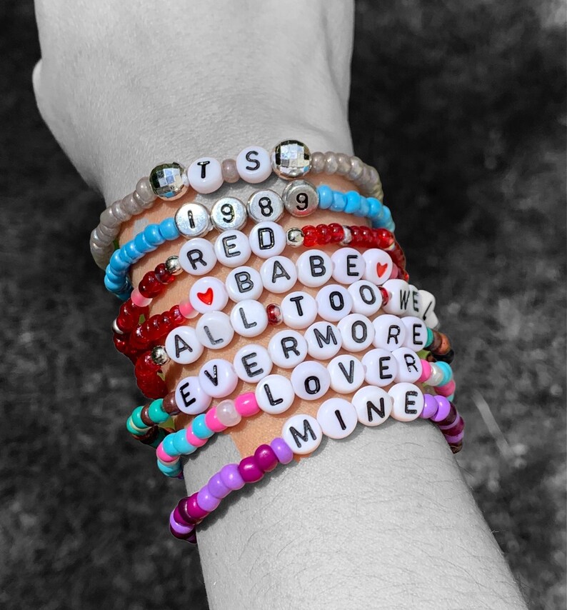 May include: A stack of colourful beaded bracelets with white letter beads spelling out the words "TS 1989 RED BABE TOO WE ALL ERMORE LOVER MINE".