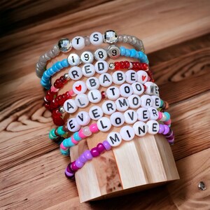May include: A stack of colourful beaded bracelets with white letter beads spelling out the words "TS 1989 RED BABE ALL TOO WE EVERMORE LOVER MINE".