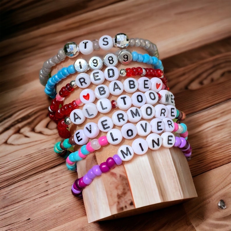 May include: A stack of colourful beaded bracelets with white letter beads spelling out the words "TS 1989 RED BABE ALL TOO WE EVERMORE LOVER MINE".