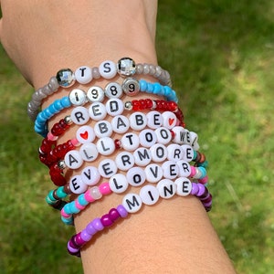 May include: A stack of colourful beaded bracelets with letter beads spelling out the words "TS 1989 RED BABE ALL TOO WE EVERMORE LOVER MINE".