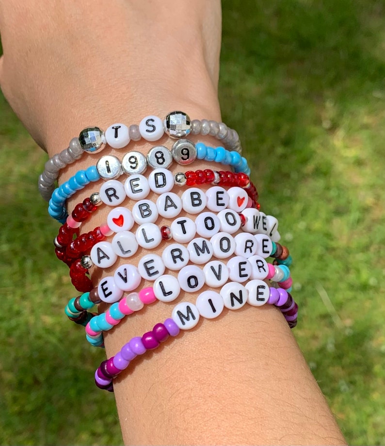 May include: A stack of colourful beaded bracelets with letter beads spelling out the words "TS 1989 RED BABE ALL TOO WE EVERMORE LOVER MINE".