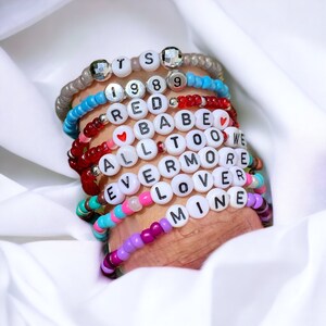 May include: A stack of colourful beaded bracelets with white letter beads spelling out the words "ITS 1989 RED BABE ALL TOO WE EVERMORE LOVER MINE".