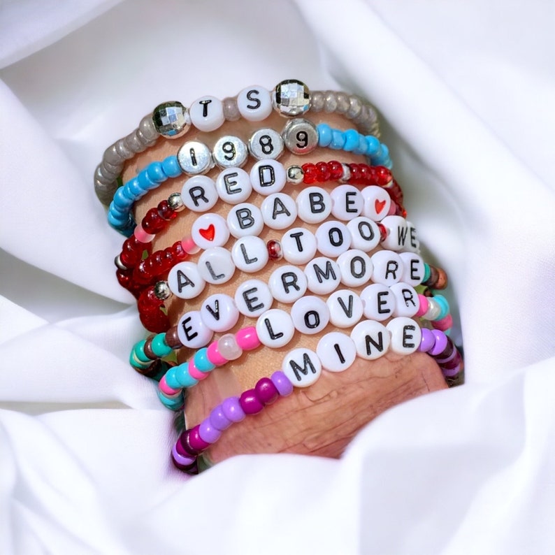 May include: A stack of colourful beaded bracelets with white letter beads spelling out the words "ITS 1989 RED BABE ALL TOO WE EVERMORE LOVER MINE".