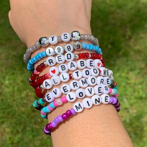 May include: A stack of colourful beaded bracelets with words like "TS", "1989", "RED", "BABE", "ALL TOO WELL", "EVERMORE", "LOVER", and "MINE".