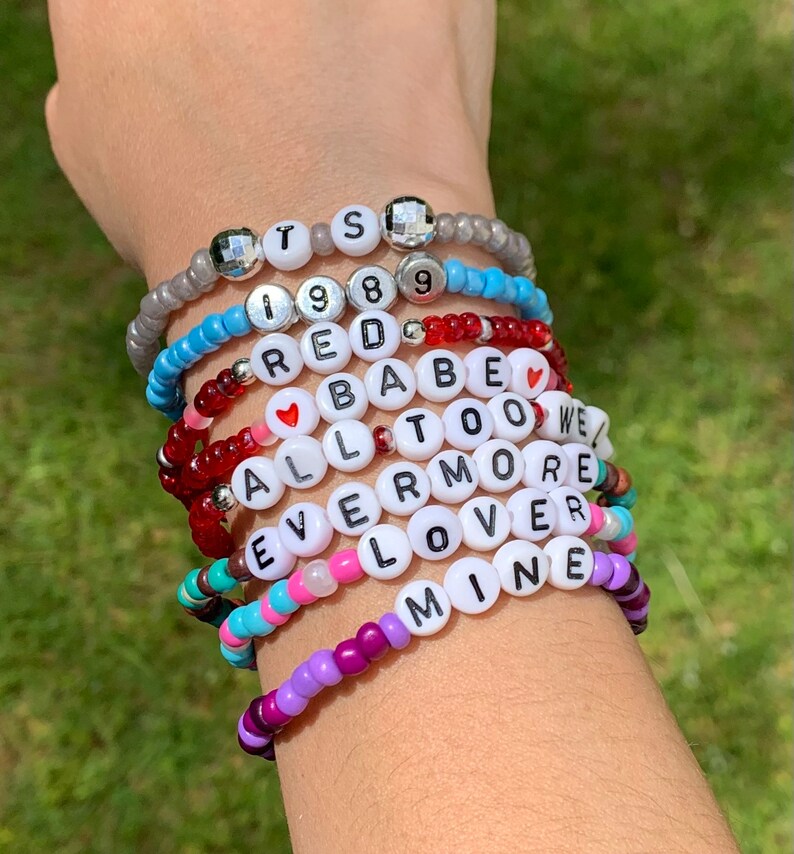 May include: A stack of colourful beaded bracelets with words like "TS", "1989", "RED", "BABE", "ALL TOO WELL", "EVERMORE", "LOVER", and "MINE".