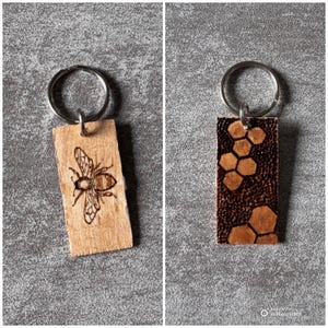 May include: Two wooden keychains with metal rings. One keychain features a detailed etched bee design, while the other showcases a honeycomb pattern. Both are rectangular and made of light-colored wood.