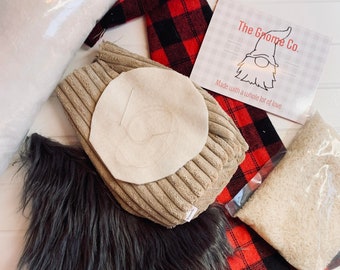 Puede incluir: Un kit de manualidades de gnomo con tela de cuadros rojos y negros, relleno blanco, un gorro de gnomo de pana marrón, un círculo de fieltro blanco y una nariz de fieltro blanco. El kit también incluye una tarjeta impresa con el texto "The Gnome Co. Made with a whole lot of love."