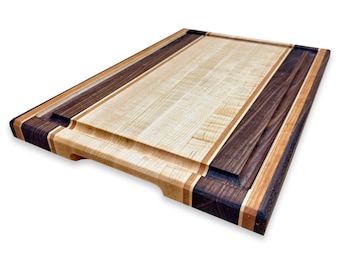 May include: A large, rectangular cutting board made of maple and walnut wood. The board has a raised edge and two handles. The wood grain is visible and the board is in good condition.