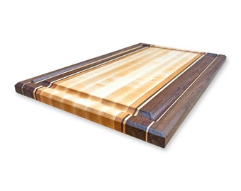 May include: A large wooden cutting board with a light brown maple surface and a dark brown walnut border. The board has a raised edge and a beveled edge.