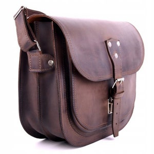 May include: A brown leather messenger bag with a flap closure and a buckle. The bag has a long adjustable strap and a small pocket on the front.