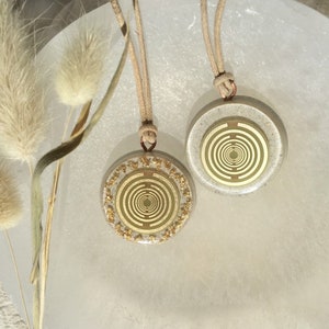 Puede incluir: Dos colgantes circulares dorados y blancos con un diseño en espiral, colgando de cordones beige. Los colgantes están sobre un fondo blanco.