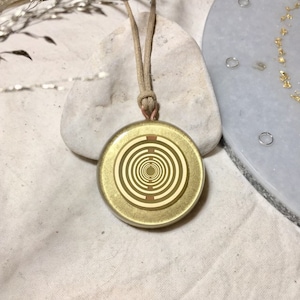 Könnte beinhalten: Eine goldene Halskette mit einem runden Anhänger, der konzentrische Kreise in hellbrauner Farbe zeigt. Der Anhänger befindet sich an einer hellbraunen Schnur.