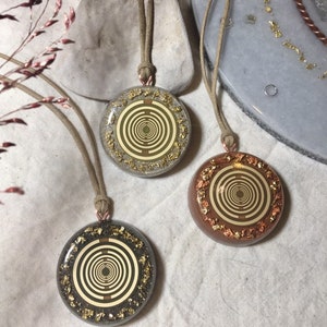 Puede incluir: Tres colgantes redondos con un diseño en espiral dorado y marrón sobre un fondo blanco. Los colgantes están en cordones marrones y cuelgan de una superficie blanca.