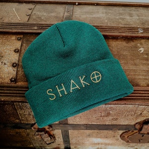 Könnte beinhalten: Eine dunkelgrüne Mütze mit dem Wort "SHAKO" und einem Kreis mit einem Kreuz darin, das in goldener Farbe gestickt ist. Die Mütze liegt auf einer Holzkiste.