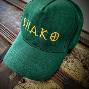May include: A green baseball cap with a yellow embroidered logo that says "SHAKO" with a yellow circle and cross inside the "O".