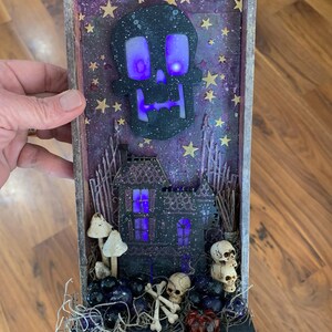 May include: A Halloween shadow box featuring a skull, a haunted house, and other decorative items. The skull and house have purple lights. The box includes miniature skulls, bones, and a small pumpkin. The background is a starry purple.