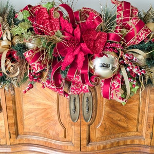 May include: A festive Christmas garland with red poinsettias, gold and red patterned ribbons, and gold ornaments. The garland is arranged on top of a wooden cabinet with brass hardware. The text "Alchemy Altered Design" is visible in the bottom right corner.