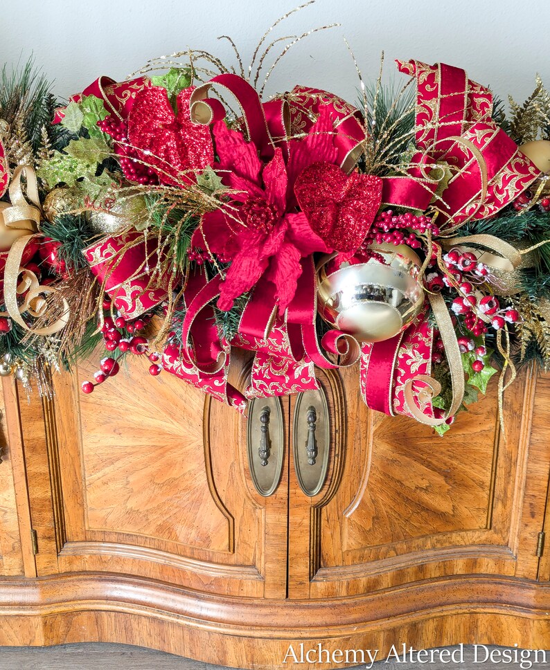 May include: A festive Christmas garland with red poinsettias, gold and red patterned ribbons, and gold ornaments. The garland is arranged on top of a wooden cabinet with brass hardware. The text "Alchemy Altered Design" is visible in the bottom right corner.
