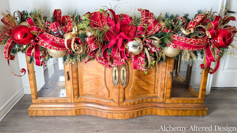 May include: A festive Christmas garland with red and gold ornaments, ribbons, and greenery, displayed atop a wooden cabinet. The garland features large red baubles, gold accents, and decorative bows, creating a holiday theme.