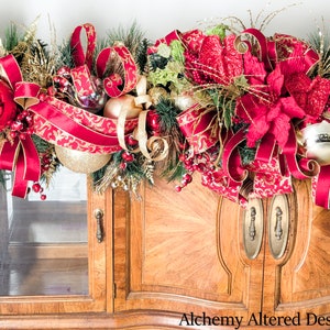 May include: A festive Christmas garland with red and gold accents. The garland features large red ornaments, gold ribbons, and faux greenery. The garland is draped over a wooden cabinet with glass doors. The text "Alchemy Altered Design" is visible.