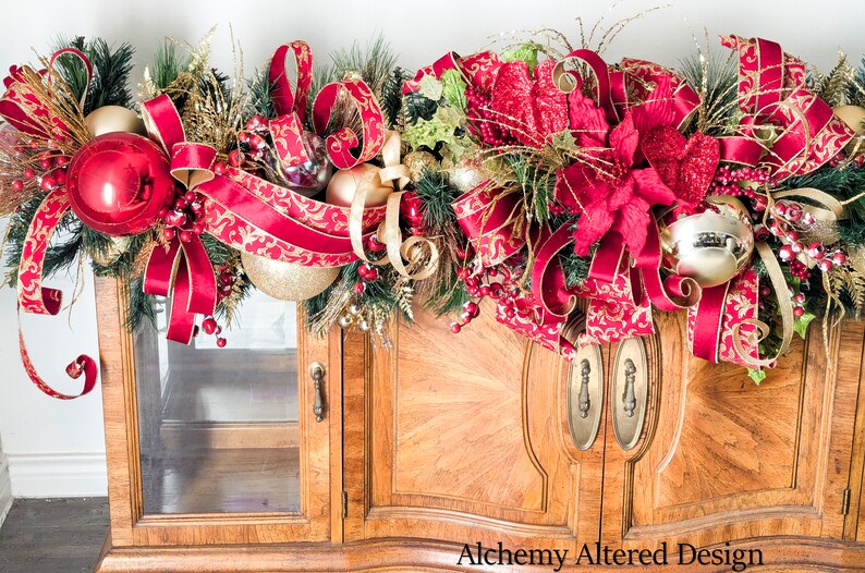 May include: A festive Christmas garland with red and gold accents. The garland features large red ornaments, gold ribbons, and faux greenery. The garland is draped over a wooden cabinet with glass doors. The text "Alchemy Altered Design" is visible.