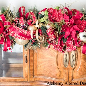 May include: A festive Christmas garland with red and gold accents. The garland features large red ornaments, gold ribbons, and faux greenery. The design includes red bows and gold-patterned ribbon, creating a luxurious holiday decoration. The garland is displayed on a wooden cabinet.