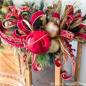 May include: A festive Christmas decoration featuring a vibrant red and gold color scheme. The arrangement includes large red ornaments, gold-colored baubles, and lush greenery, accented with ornate red and gold ribbons. The design is set against a wooden cabinet.