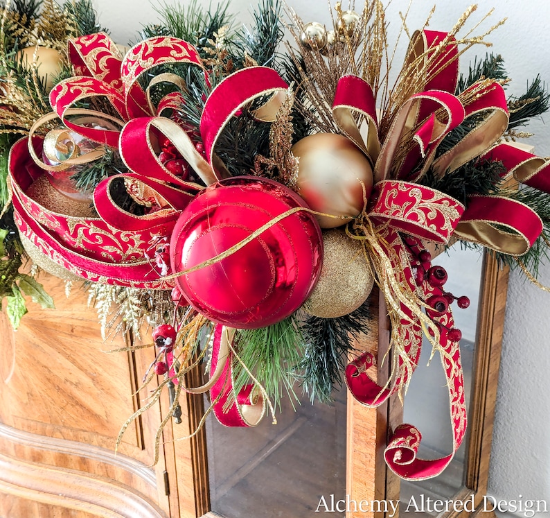 May include: A festive Christmas decoration featuring a vibrant red and gold color scheme. The arrangement includes large red ornaments, gold-colored baubles, and lush greenery, accented with ornate red and gold ribbons. The design is set against a wooden cabinet.