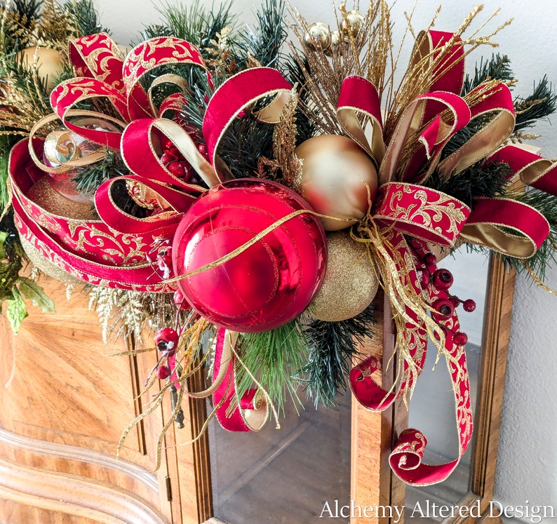 May include: A festive Christmas decoration featuring a vibrant red and gold color scheme. The arrangement includes large red ornaments, gold-colored baubles, and lush greenery, accented with ornate red and gold ribbons. The design is set against a wooden cabinet.