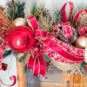 May include: A festive Christmas garland featuring a large red ornament, gold and red ribbons, and gold accents. The garland includes faux berries, clear and gold ornaments, and evergreen branches, creating a holiday display.