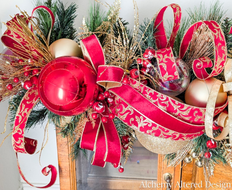 May include: A festive Christmas garland featuring a large red ornament, gold and red ribbons, and gold accents. The garland includes faux berries, clear and gold ornaments, and evergreen branches, creating a holiday display.