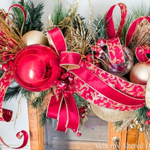 May include: A festive Christmas garland featuring a large red ornament, gold and red ribbons, and gold accents. The garland includes faux evergreen branches, gold berries, and clear and gold ornaments, creating a holiday display.