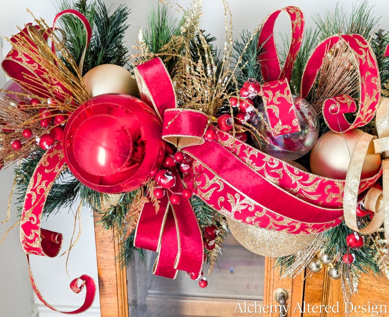 May include: A festive Christmas garland featuring a large red ornament, gold and red ribbons, and gold accents. The garland includes faux evergreen branches, gold berries, and clear and gold ornaments, creating a holiday display.