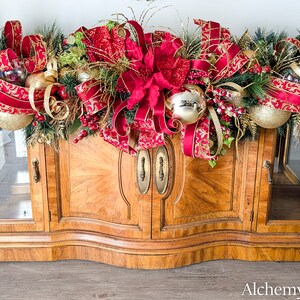 May include: A festive Christmas garland with red and gold ornaments, ribbons, and greenery, displayed atop a wooden cabinet. The garland features large red baubles, gold accents, and decorative bows, creating a holiday display.