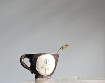 Könnte beinhalten: Eine kleine braune Keramiktasse mit einem weißen Blumenmuster und einem grünen Stiel mit einer braunen Knospe, die aus der Tasse ragt.
