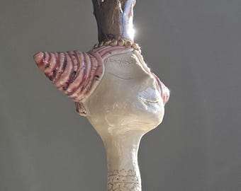 May include: A ceramic sculpture of a stylized head wearing a crown. The head is off-white with pink accents on the ear and around the crown. The crown is brown and gold. The sculpture has a long neck and a decorative base.