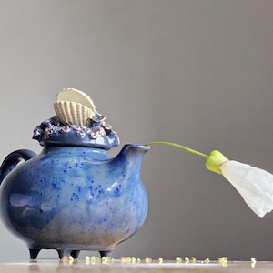 May include: A blue ceramic teapot with a seashell lid and a white flower on a stem. The teapot has a speckled blue glaze and three small feet.