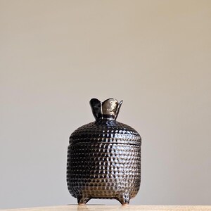 Può includere: Vaso in ceramica nera con superficie testurizzata e coperchio a forma di fiore. Il vaso ha tre piccoli piedini.